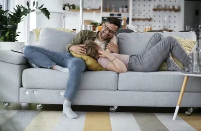 A smiling couple is relaxing intimately on a gray couch in a living room.