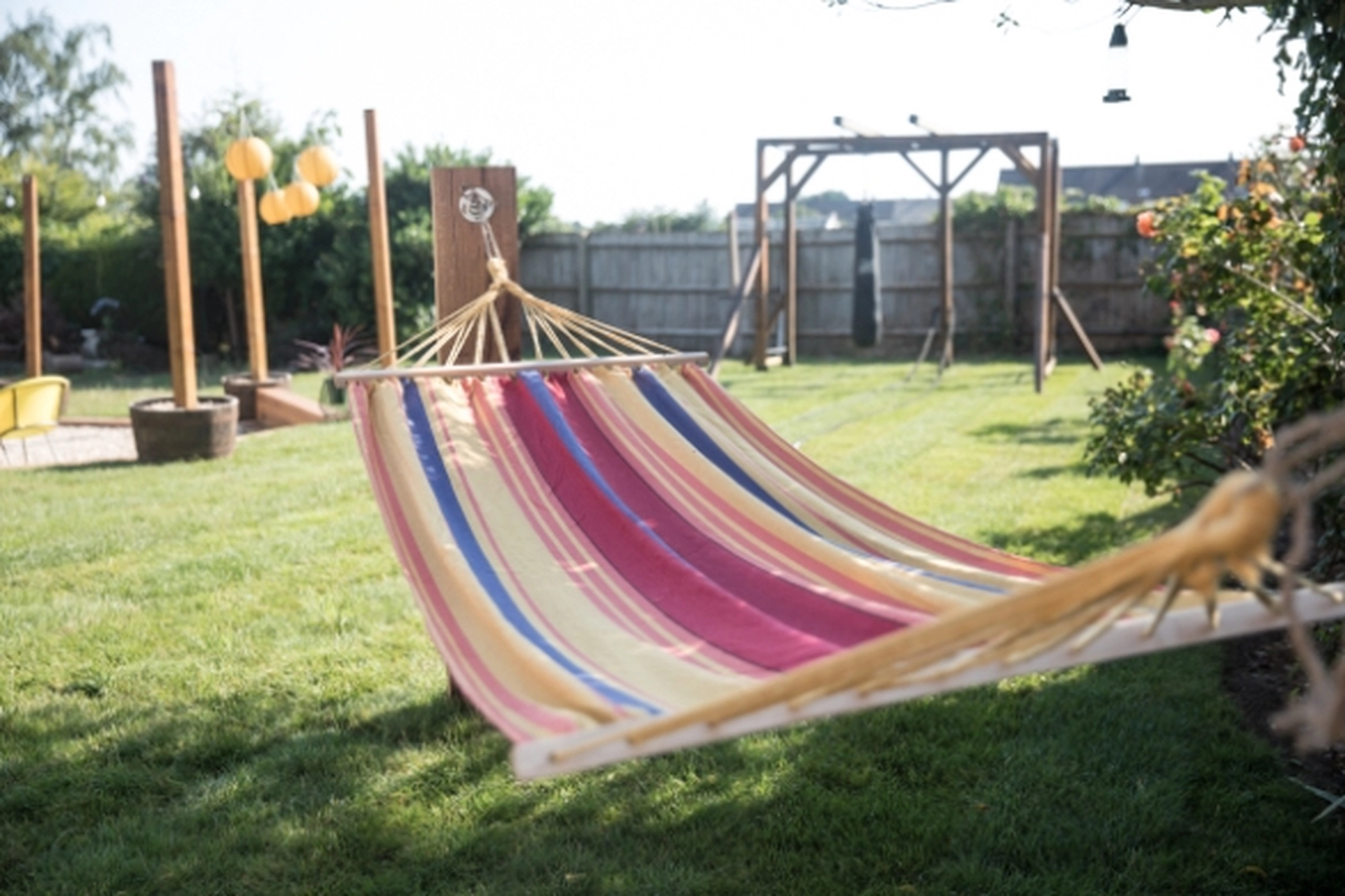 Colorful striped hammock hanging over a grassy backyard, with a wooden structure and decorative outdoor lights in the background on a sunny day.